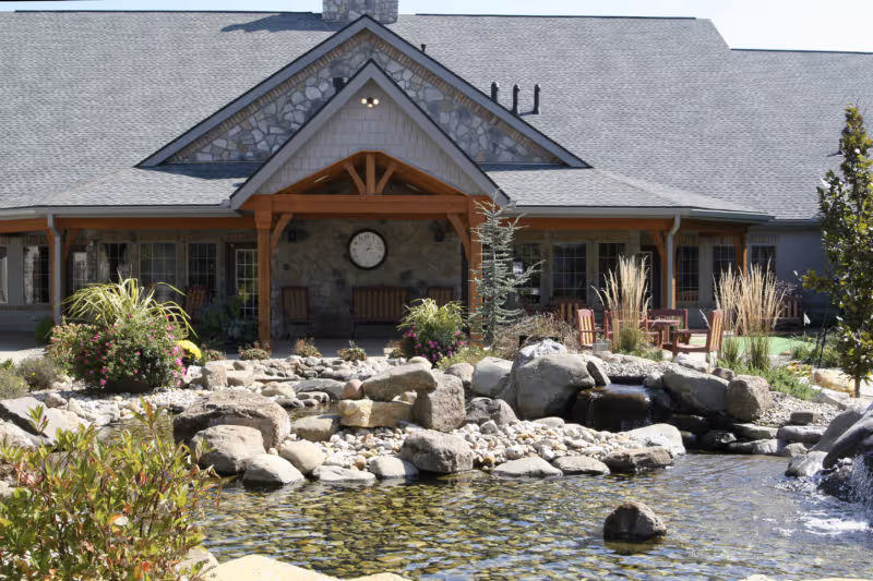 Stone-faced building entrance with a covered wooden porch, outdoor seating, landscaped rocks and a pond in the foreground.