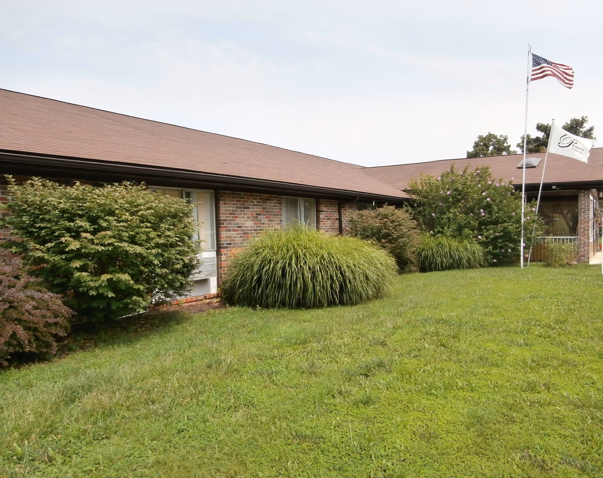 Exterior view of a single-story brick building with a brown roof, surrounded by green grass and various bushes. Two flagpoles are visible, one with an American flag and the other with a white flag featuring a logo. The sky is overcast.