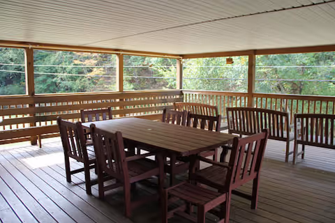 Covered wooden porch with a rectangular dining table and several wooden chairs overlooking trees.