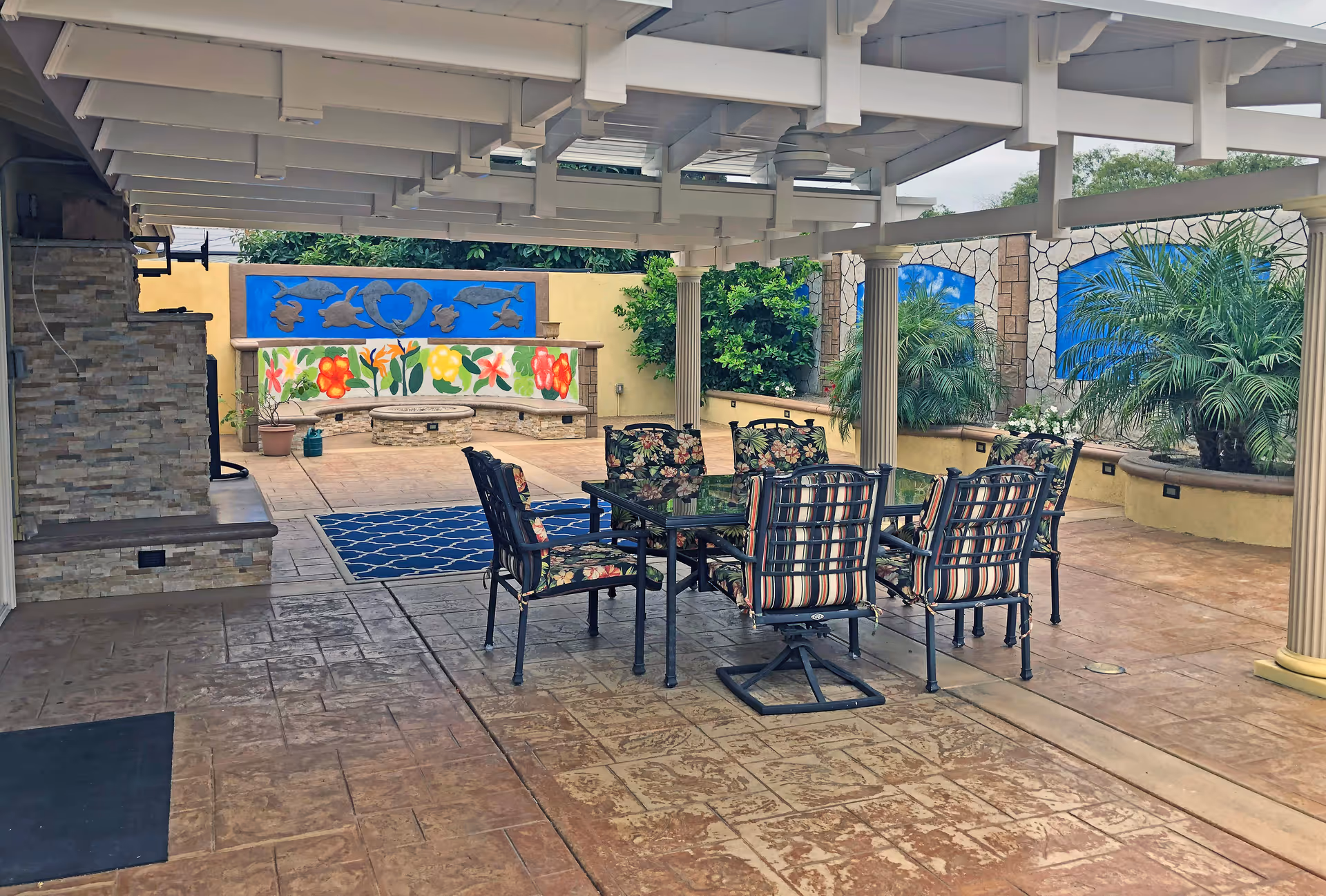 Covered outdoor patio area with a glass-top table and six cushioned chairs featuring floral and striped patterns. The patio has stone flooring and is supported by white columns. In the background, there is a curved stone bench with colorful floral murals and blue panels with sea life designs. There are also potted plants and greenery around the space.
