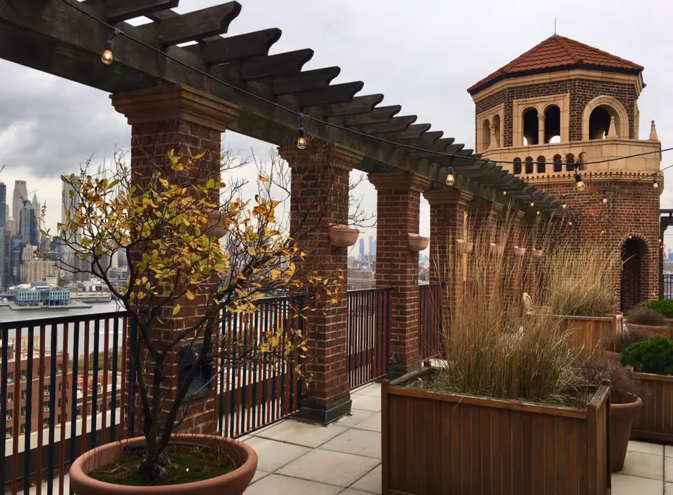Outdoor terrace with brick columns supporting a wooden pergola adorned with string lights. There are potted plants and tall grasses along the railing, and a brick tower structure with arched openings and a red-tiled roof. In the background, a city skyline is visible under a cloudy sky.