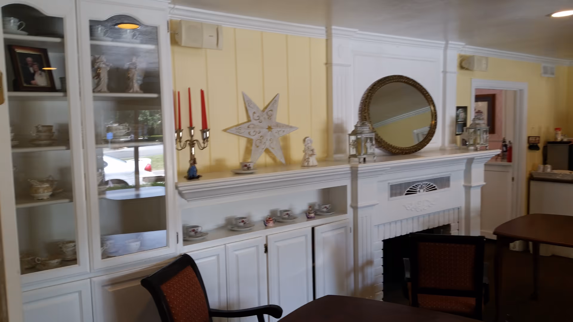 Interior view of a cozy room with yellow walls featuring a white built-in cabinet displaying teacups and decorative items, a white fireplace with a round mirror above it, and a dining table with chairs.