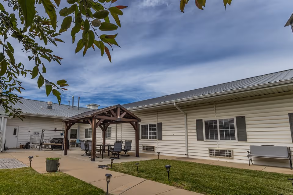 Outdoor patio area at Bailey Pointe at Plattsmouth featuring a wooden pergola with a table and chairs underneath, a bench along the building wall, green grass, and a partly cloudy sky.