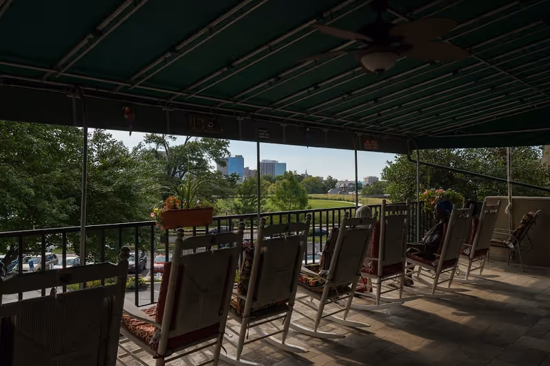 Covered outdoor patio area with a row of rocking chairs facing a scenic view of trees, a grassy area, and city buildings in the distance. A person is sitting in one of the rocking chairs on the right side. The patio has a tiled floor and a green canopy overhead.