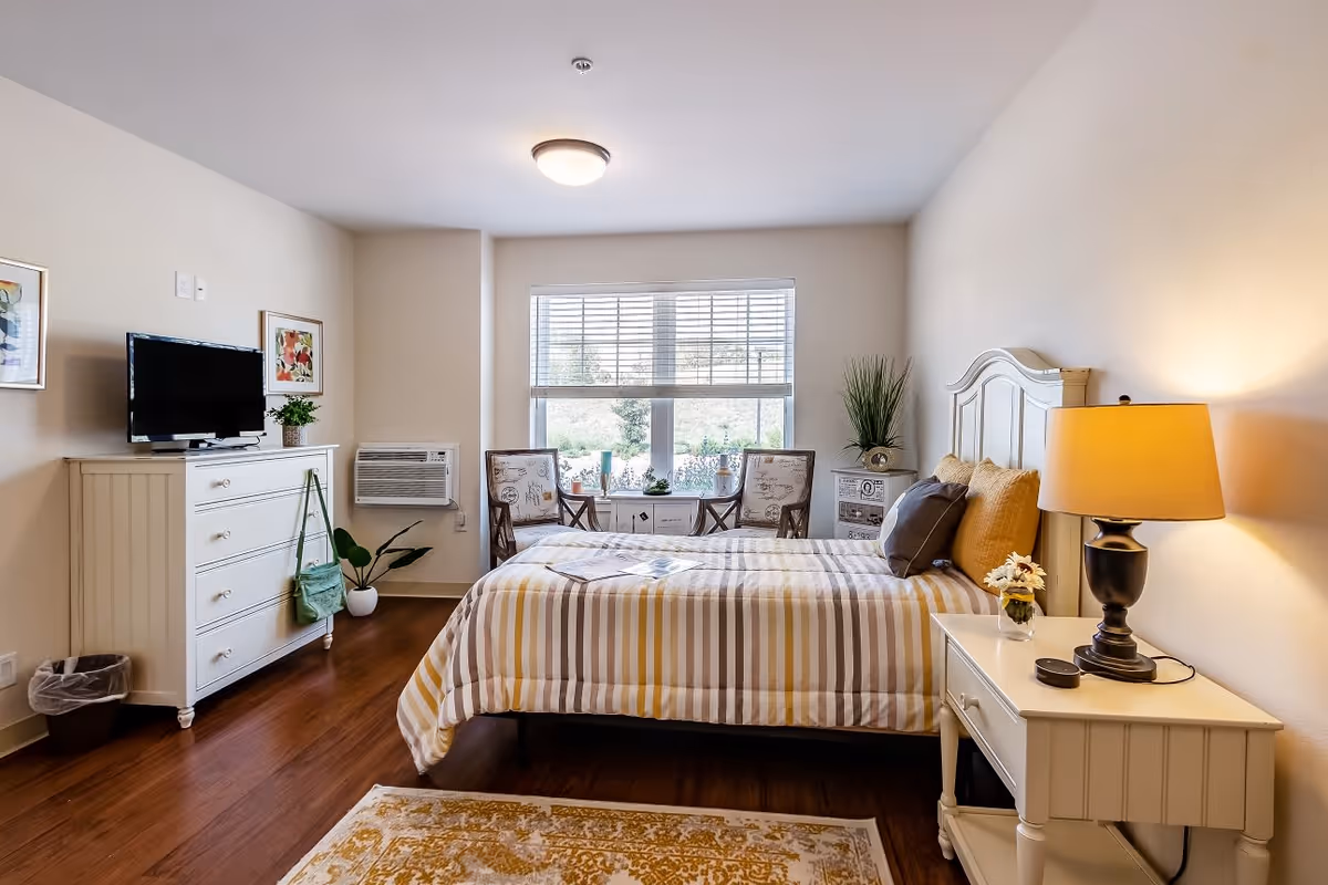 A bright and cozy bedroom with a large window letting in natural light. The room features a bed with striped bedding and multiple pillows, a nightstand with a lamp and a small flower vase, a dresser with a TV on top, two chairs with a small table between them near the window, and wooden flooring with a patterned rug.