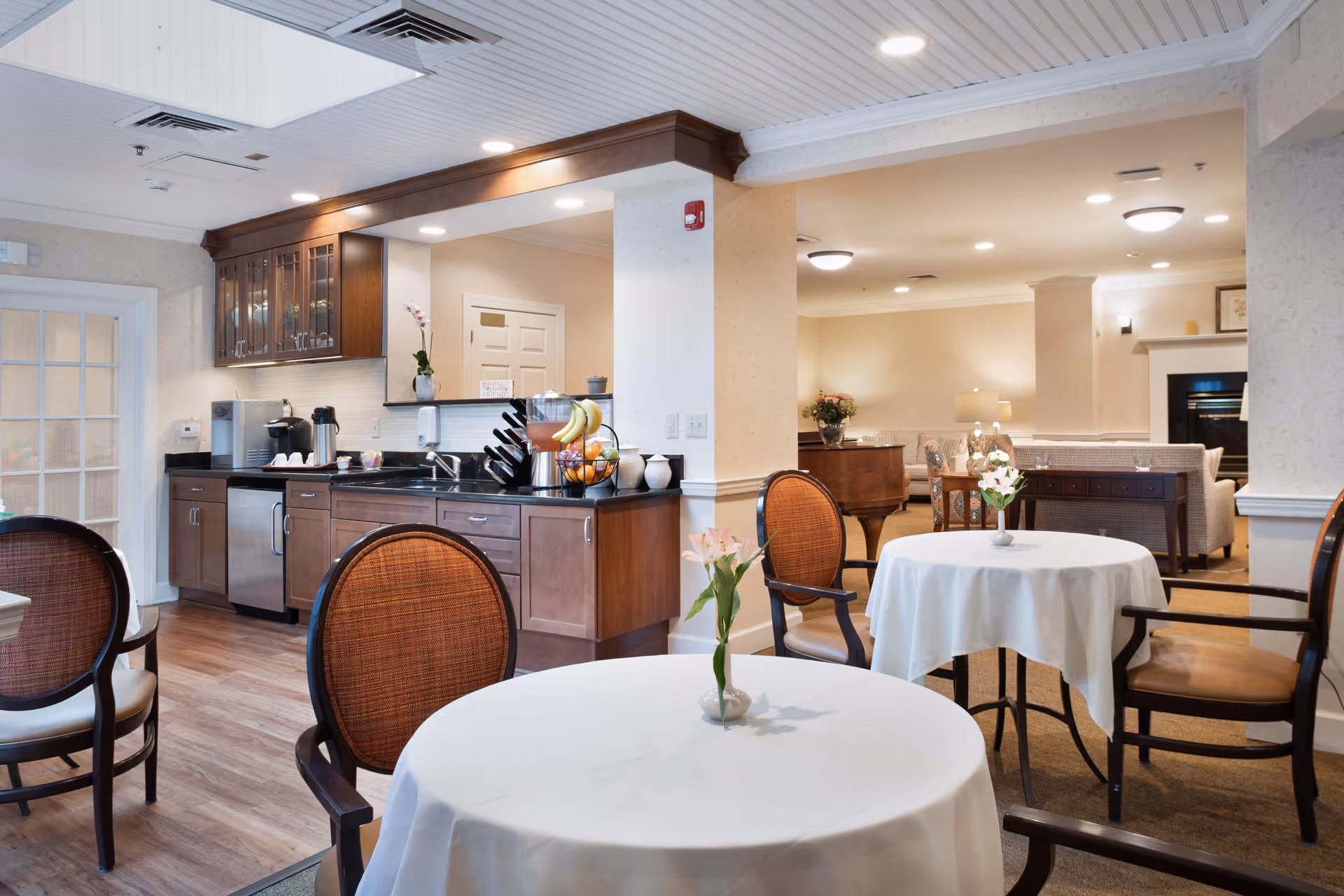 A cozy dining area in a senior living facility with round tables covered in white tablecloths, each adorned with a small vase of flowers. The room features wooden chairs with cushioned seats and backs. In the background, there is a kitchenette with dark wood cabinets, a coffee machine, and a fruit basket. Beyond the dining area, a comfortable living room space with sofas, a piano, and a fireplace is visible.