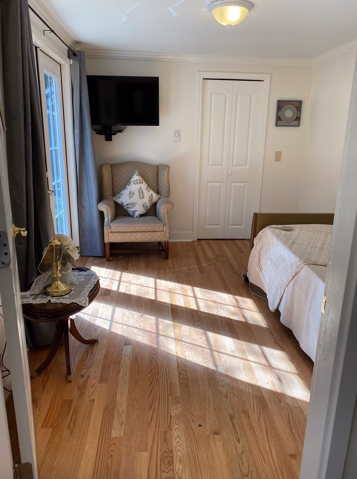 A bright bedroom with wooden flooring, a bed with white bedding on the right, a beige armchair with a patterned cushion against the wall, a wall-mounted flat screen TV above the chair, a small round wooden side table with a brass lamp and lace doily near the door, and sunlight streaming through a window with gray curtains on the left.