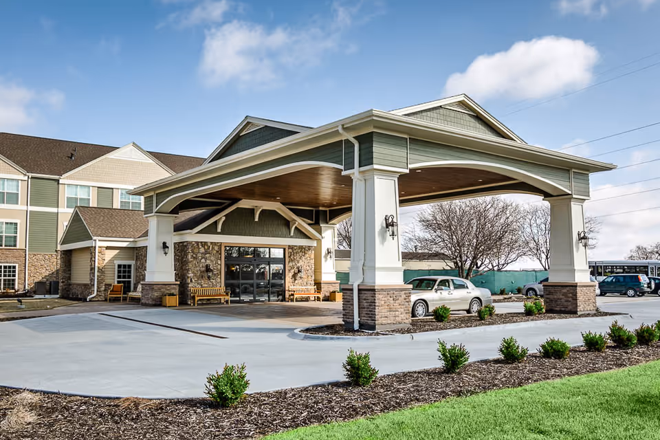 Entrance of Remington Heights Retirement Community featuring a covered drop-off area with large white pillars and a stone facade. There are benches near the entrance, a few parked cars, and landscaped greenery with small bushes and grass. The building has multiple windows and a pitched roof under a partly cloudy sky.