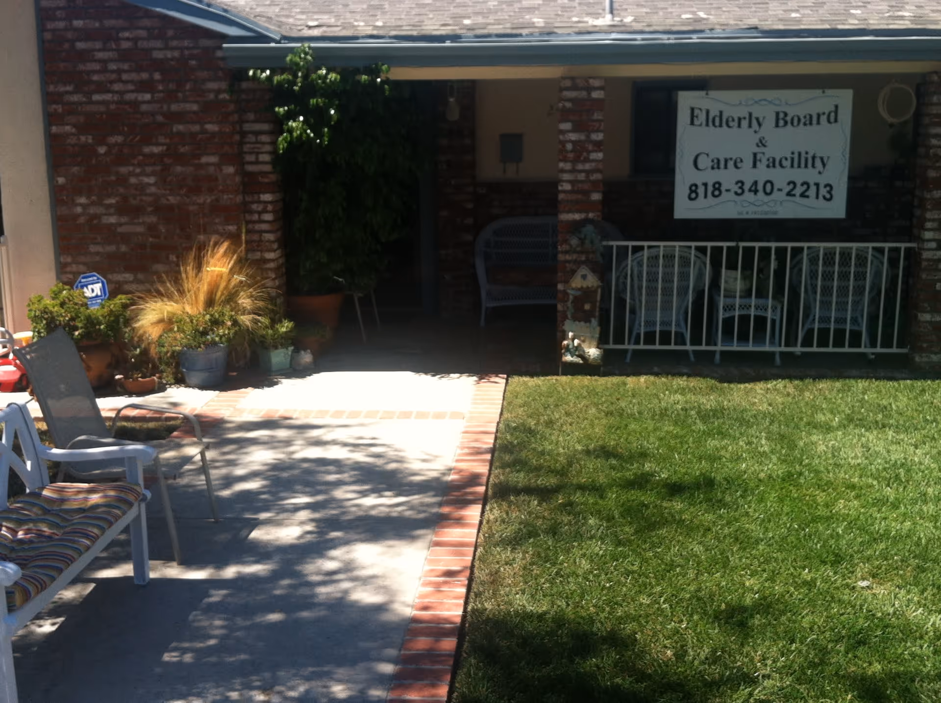 Front porch area of a brick building with a small lawn and paved walkway. There are several chairs and potted plants on the porch. A sign on the porch reads 'Elderly Board & Care Facility 818-340-2213'.