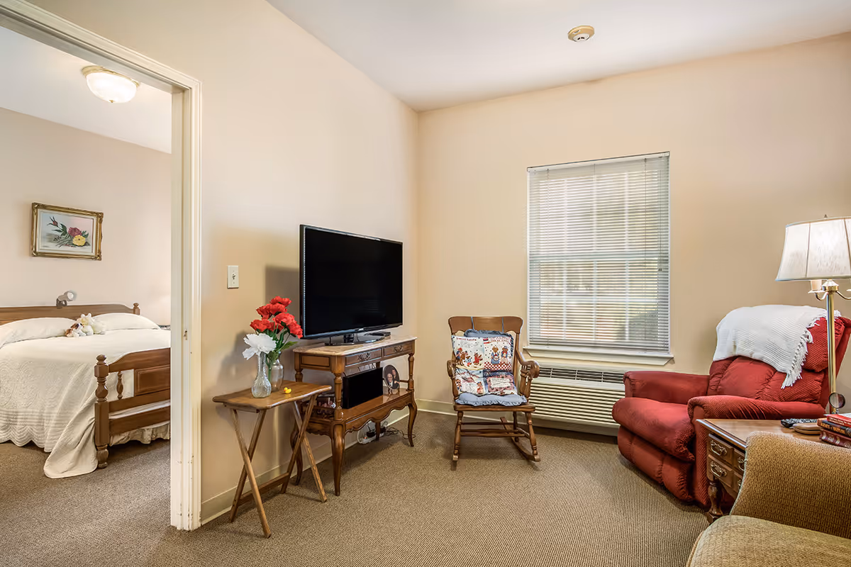 A cozy living room with a red armchair, a wooden rocking chair with a decorative cushion, a wooden side table with a vase of red and white flowers, and a flat-screen TV on a wooden stand. A window with blinds is on the wall, and an adjacent bedroom with a bed covered in a white quilt and a framed floral painting is visible through an open doorway.