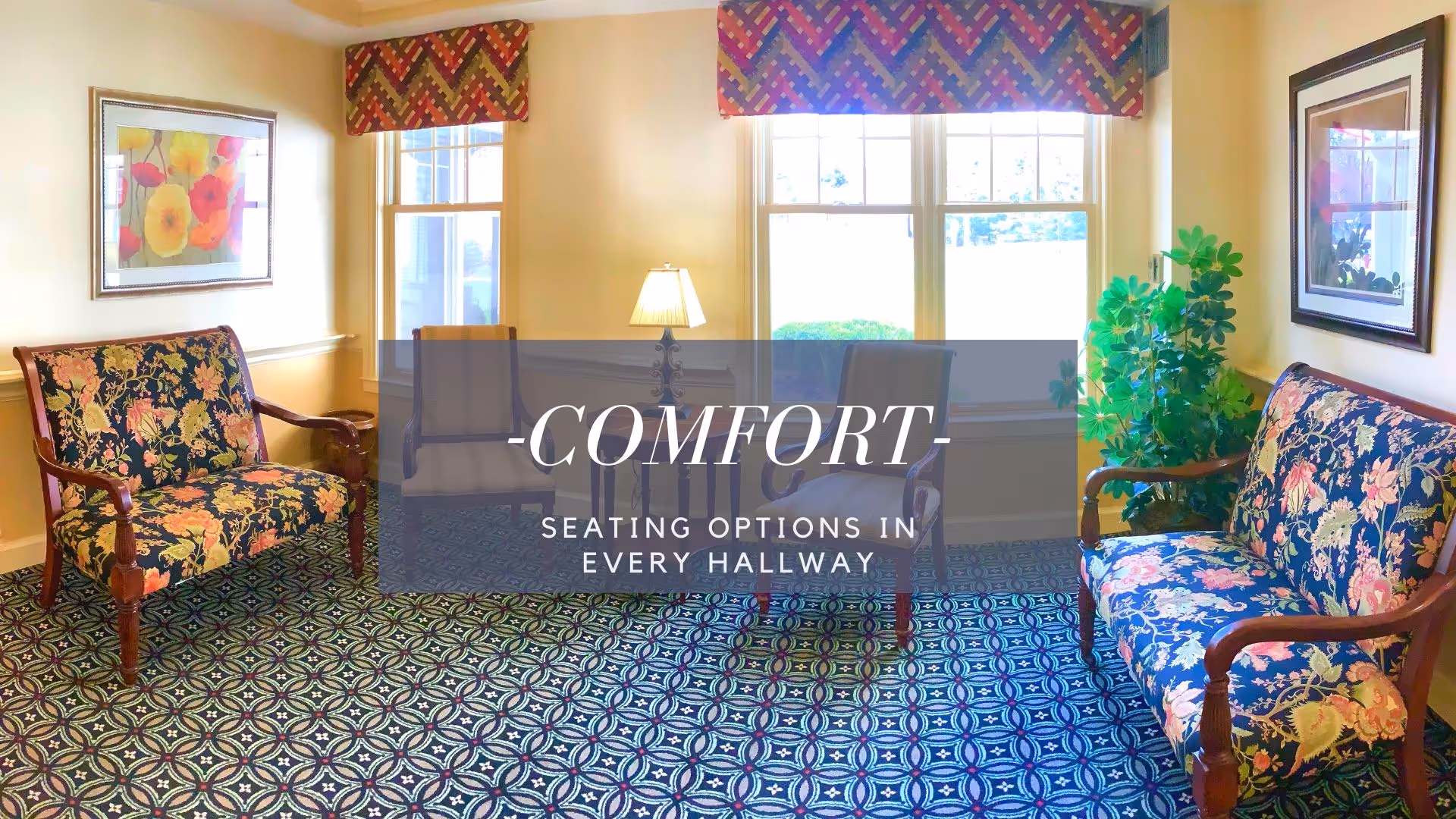 A cozy seating area in a hallway with two floral upholstered wooden armchairs, two additional chairs, a table lamp, framed floral artwork on the walls, patterned carpet, and windows with colorful valances letting in natural light.