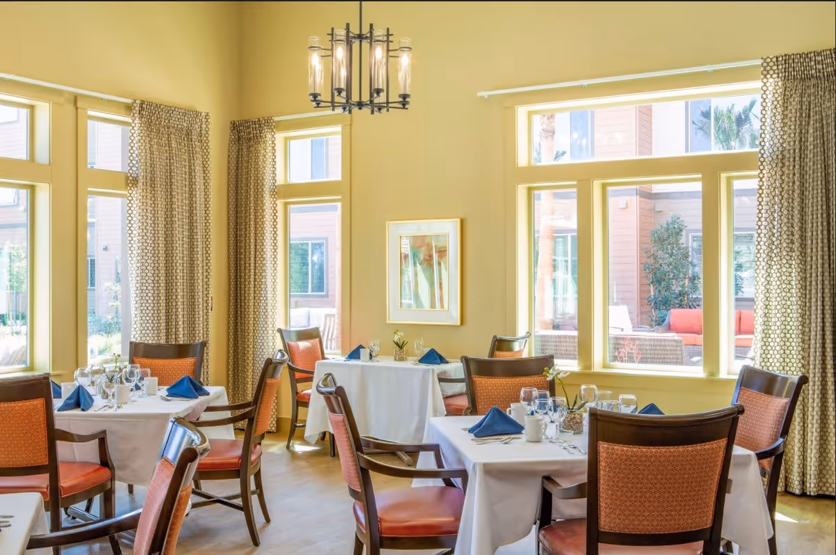 Bright dining room with several tables covered in white tablecloths, each set with blue napkins, glassware, and small flower arrangements. The room has large windows with patterned curtains allowing natural light to fill the space. Wooden chairs with orange cushions surround the tables, and a modern chandelier hangs from the ceiling.