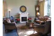 A cozy living room with a fireplace centered on the back wall, flanked by a tall grandfather clock and a floor lamp. The room features a brown leather sofa and two matching armchairs arranged around a wooden coffee table on a patterned rug. Light beige curtains cover the windows, and decorative items are placed on the mantel above the fireplace.