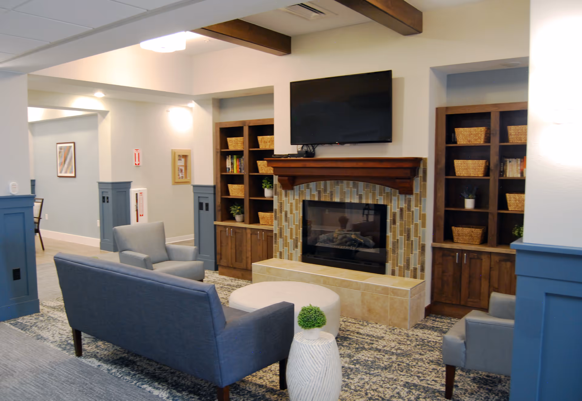 A cozy senior living common area featuring a fireplace with a tiled surround and wooden mantel, flanked by built-in wooden shelves filled with wicker baskets and plants. In front of the fireplace are two gray armchairs, a blue loveseat, a round ottoman, and a small white side table with a green plant. The room has blue wainscoting and light-colored walls with ceiling beams.