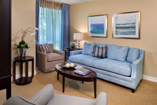 Sunlit living room featuring a light-blue sofa, two armchairs, a wooden coffee table, framed artwork, and a window with curtains.