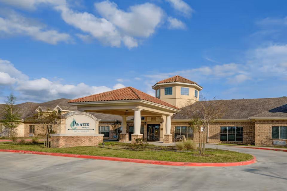 Exterior view of Proveer at Northgate facility with a covered entrance, brick walls, and a tower-like structure under a partly cloudy blue sky.