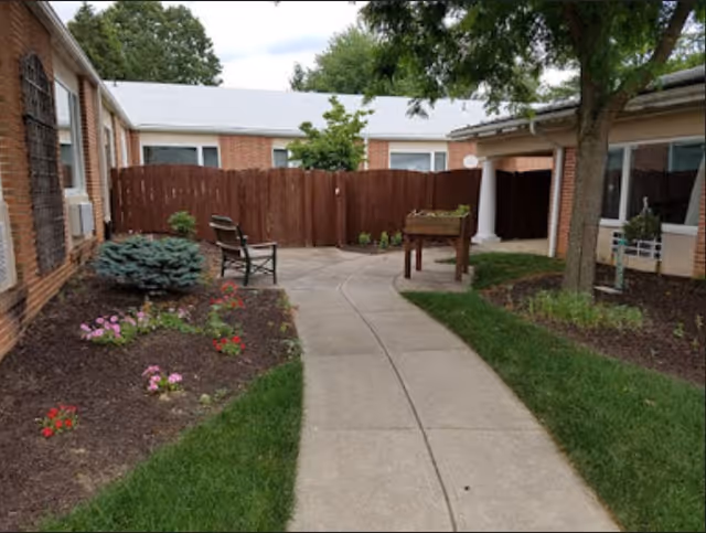 Outdoor courtyard area with a concrete pathway, green grass, flower beds with blooming flowers, a bench, a raised garden bed, and surrounding brick buildings with windows and a wooden fence.