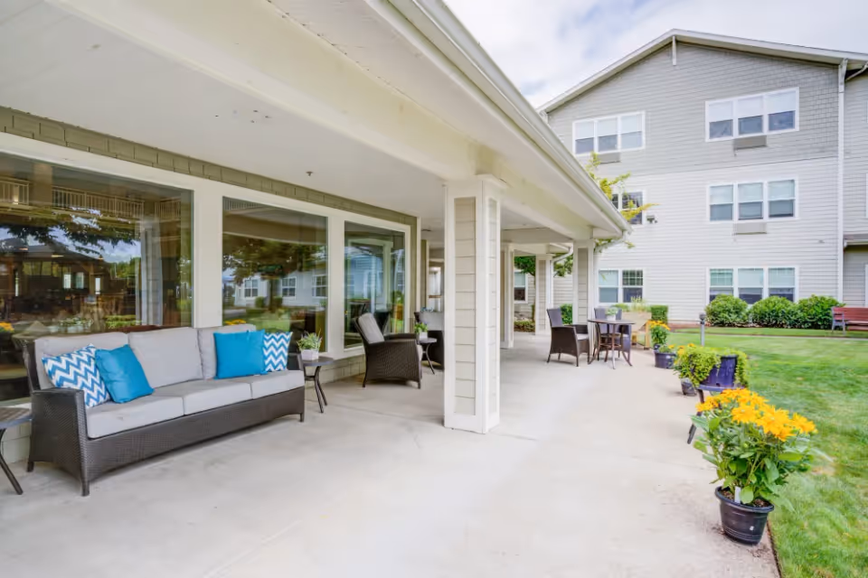 Covered outdoor patio area at Timber Pointe Senior Living with wicker seating including a sofa with blue and white cushions, chairs, small tables, and potted yellow flowers along the edge. The patio overlooks a grassy lawn and a multi-story building with multiple windows.