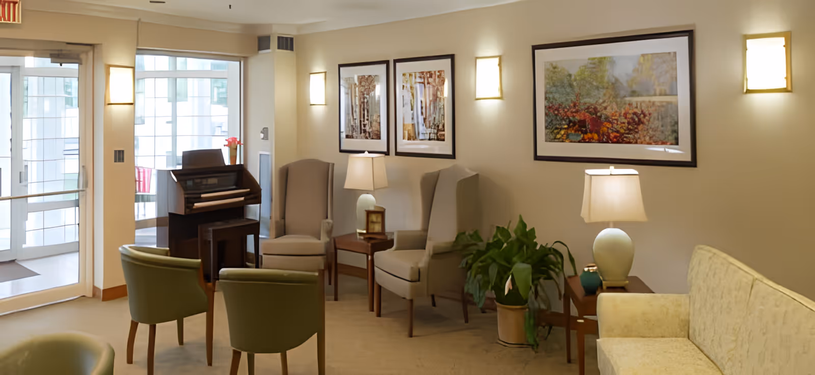 A cozy sitting area in a senior living facility with beige walls, several armchairs, a small sofa, two side tables with lamps, framed artwork on the walls, a potted plant, and a small upright piano near a glass door entrance.