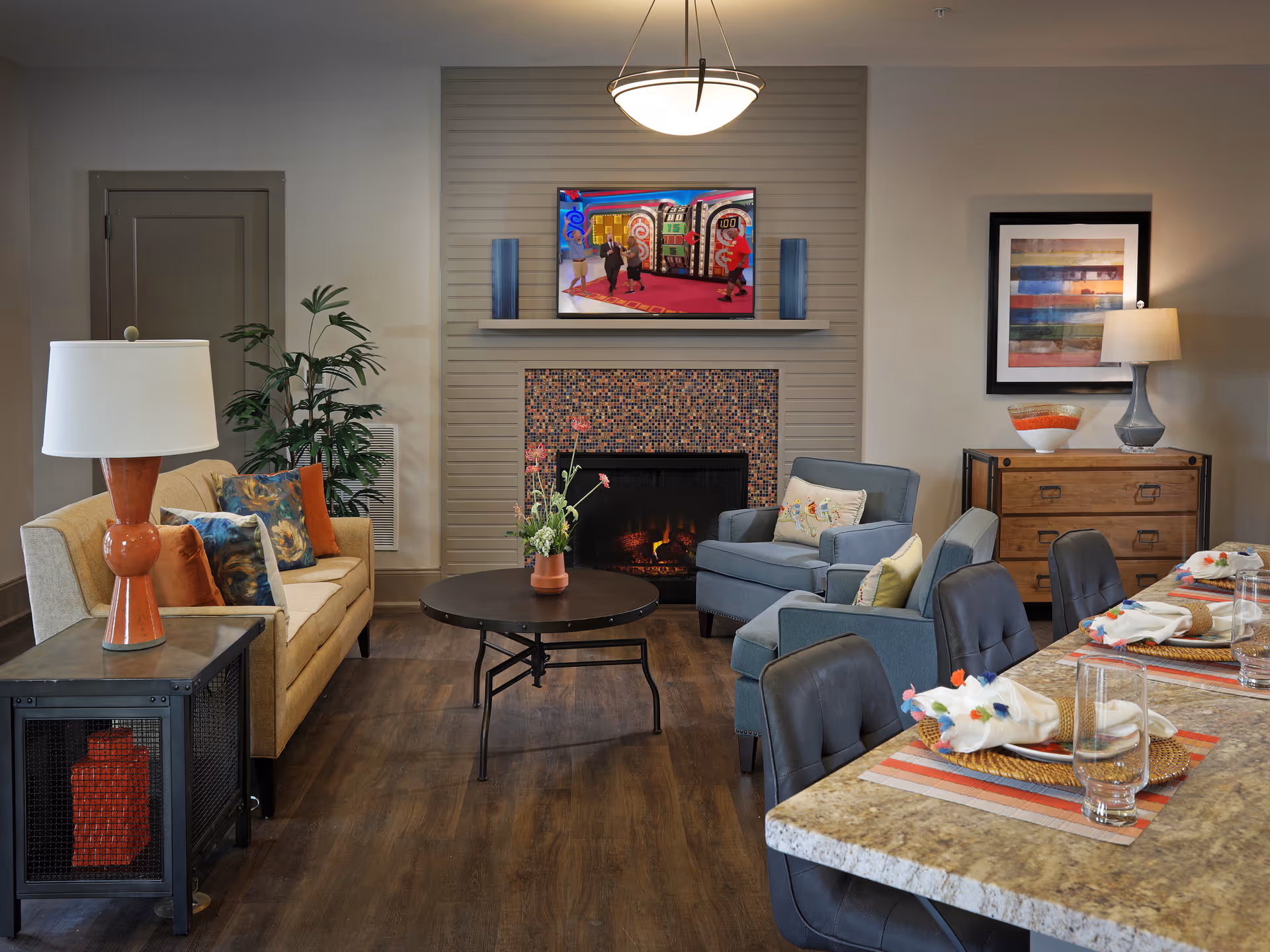 A cozy living room area with a beige sofa adorned with colorful pillows, two blue armchairs, and a round coffee table with a flower vase. A fireplace with a mosaic tile surround is centered on the back wall, above which a flat-screen TV is mounted. To the right, there is a wooden dresser with a lamp and decorative bowl, and in the foreground, a dining counter set with plates, napkins, and glasses. The room has warm lighting and wooden flooring.