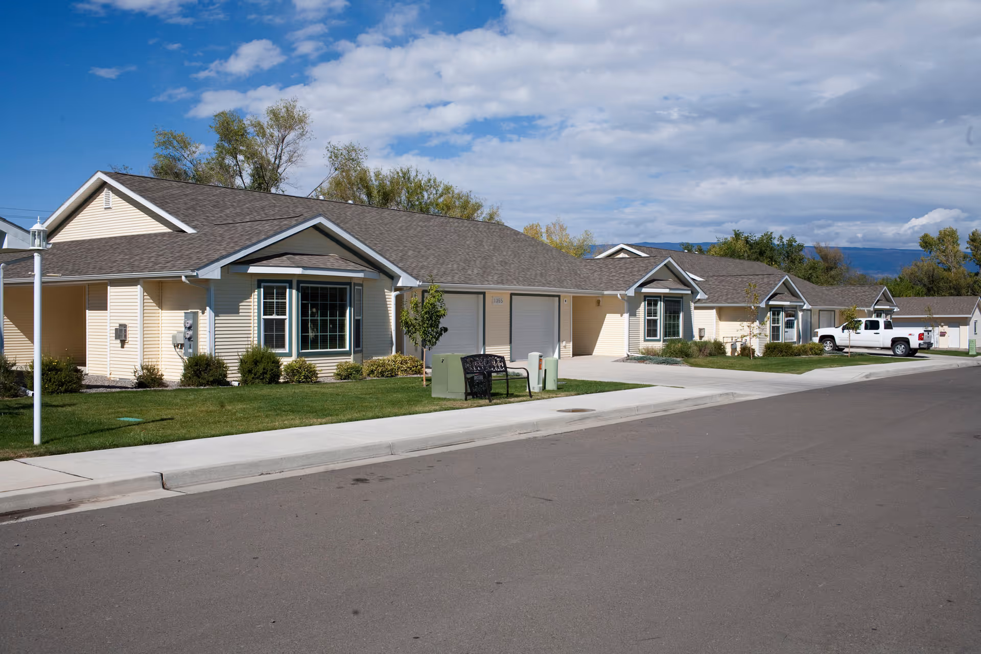 Row of single-story beige houses with attached garages, lawns, and a sidewalk along a quiet street under a partly cloudy sky.