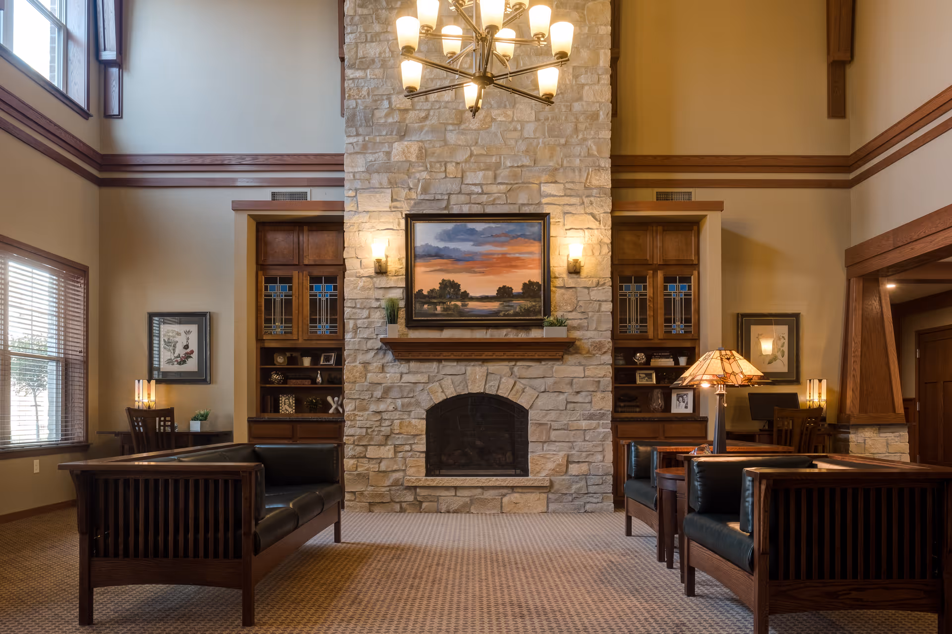 A spacious common living room with a tall stone fireplace, a framed painting above the mantel, chandelier lighting, wooden bookshelves and leather seating.