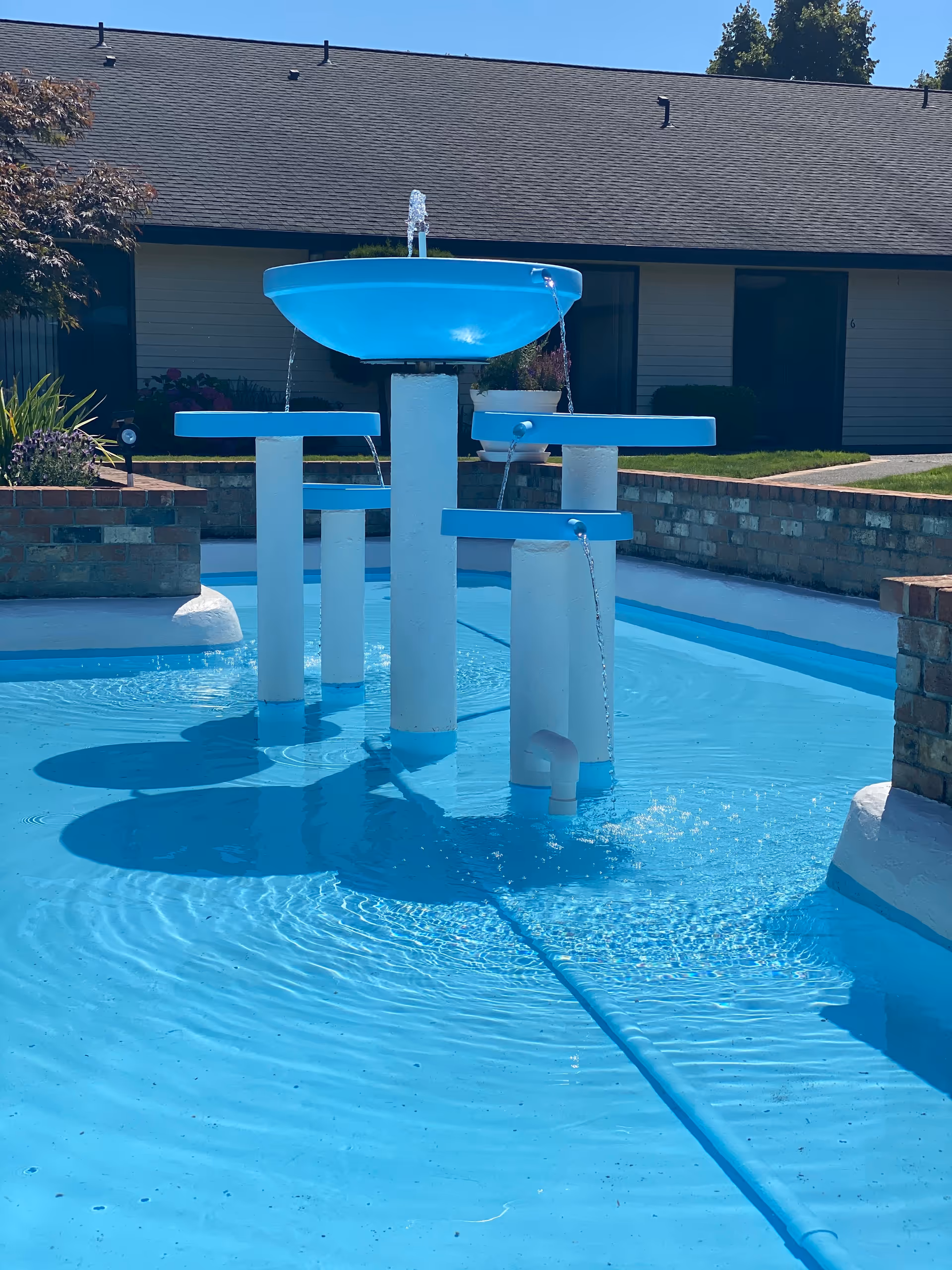 A blue and white water fountain feature in a shallow pool with water jets flowing from multiple elevated circular platforms. The background shows a single-story building with a dark roof and beige walls, surrounded by some greenery and brick edging around the pool area.