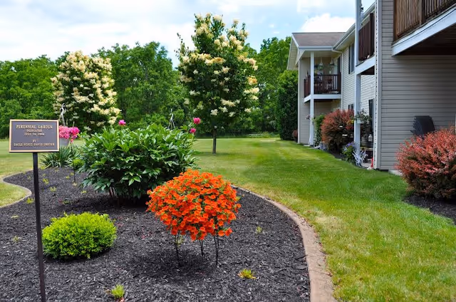 A well-maintained garden area with various flowering plants and shrubs, including a prominent cluster of orange flowers. The garden is bordered by a curved edge separating it from a green lawn. In the background, there are two-story residential buildings with balconies and some outdoor furniture. Trees and a clear sky are visible further behind the buildings.