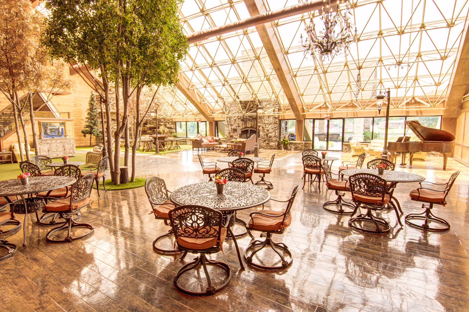 A spacious, sunlit indoor common area with a glass ceiling and large windows. The room features multiple round metal tables with cushioned chairs, several tall indoor trees, a stone fireplace, a grand piano, and comfortable seating areas. The floor is polished wood, reflecting the natural light.
