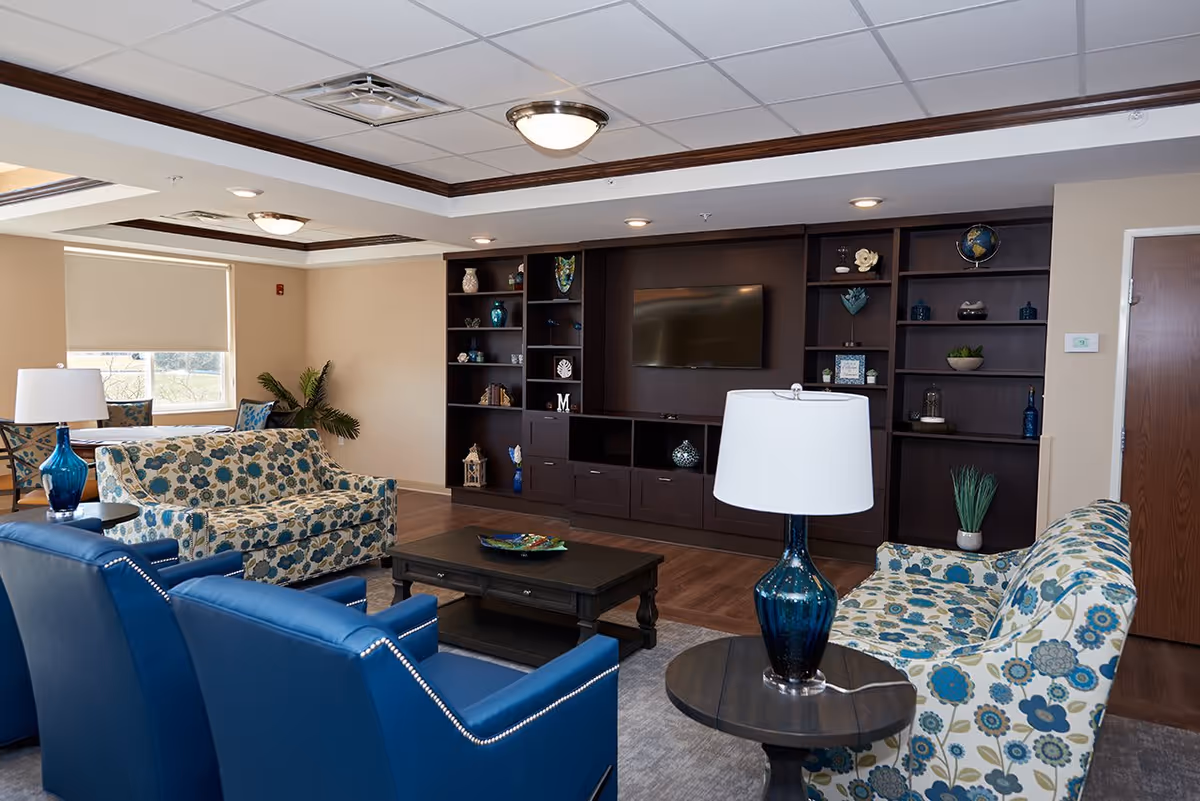 Common lounge with patterned sofas, blue armchairs, coffee table, lamps, and a wall of built-in shelving with a TV.