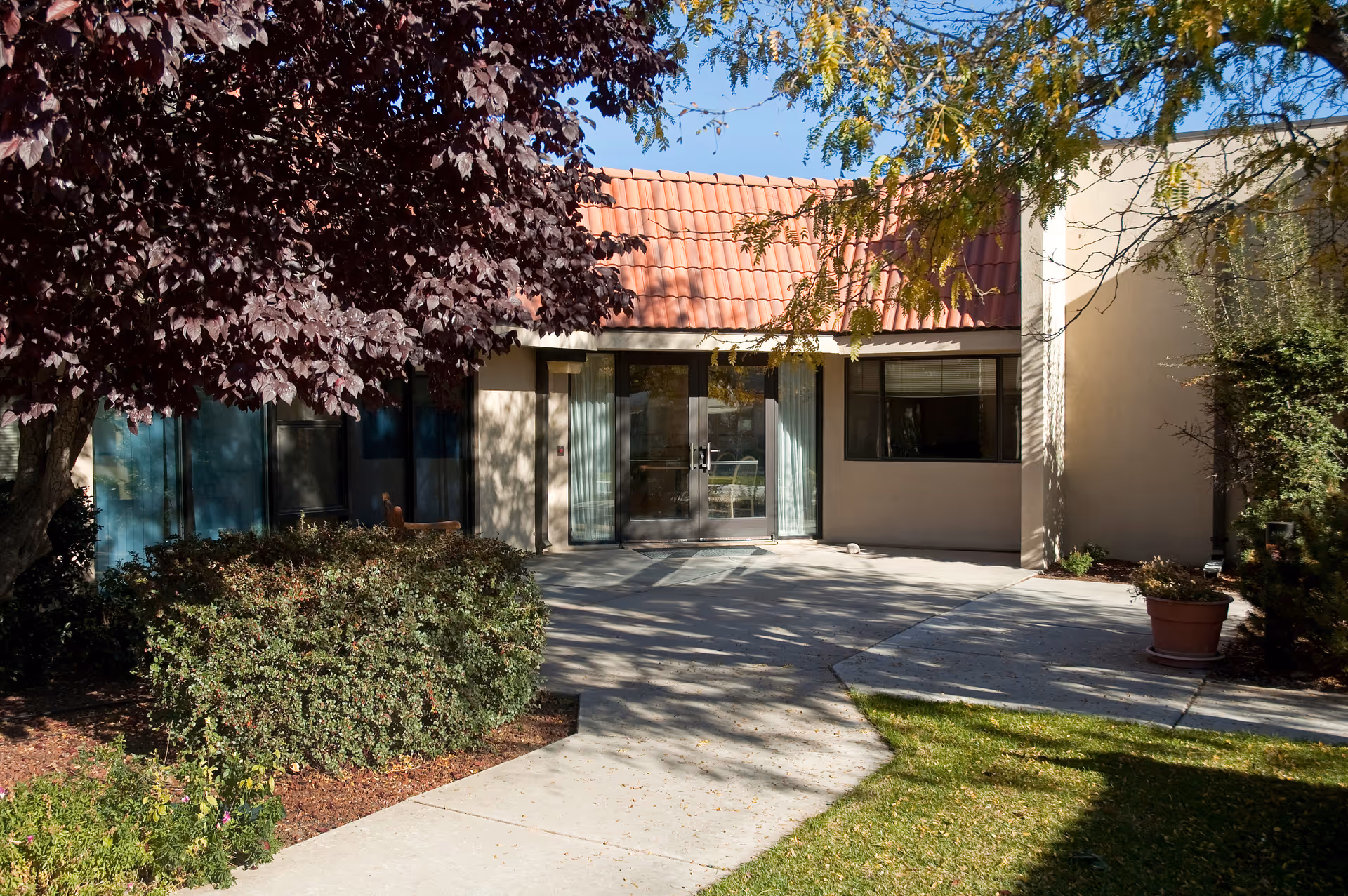 Entrance to a building with glass double doors, beige walls, and a red tiled roof. The area is surrounded by trees and bushes with a concrete walkway leading to the doors.