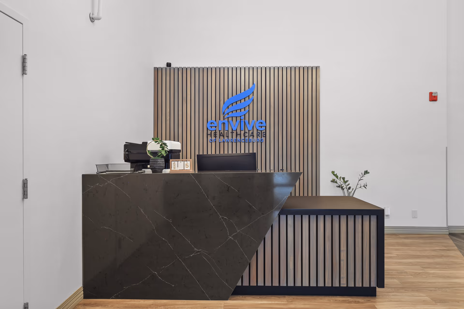 Reception desk area with a modern black marble counter and wood paneling behind it displaying the blue Envive Healthcare of Lawrenceburg logo. There is a small plant and a desk calendar on the counter, with light wood flooring and white walls surrounding the space.