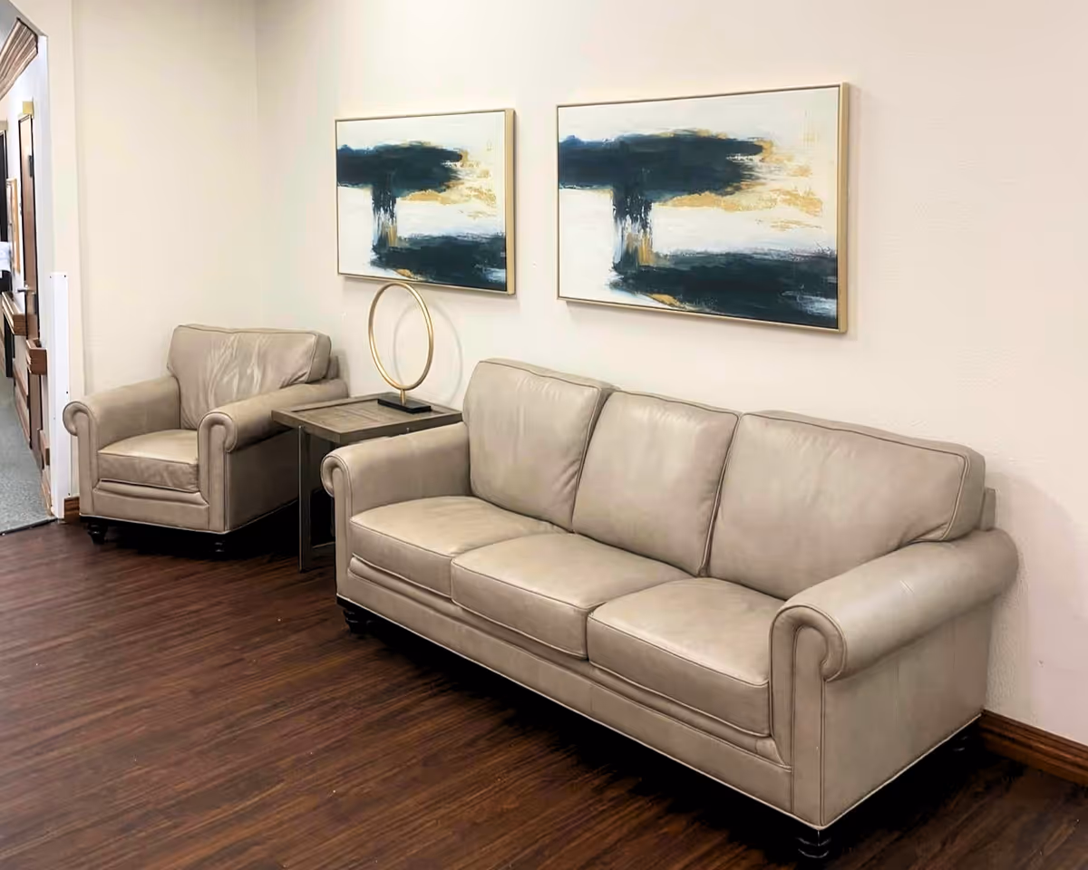 A seating area with a beige leather three-seat sofa and a matching armchair separated by a small side table with a decorative circular sculpture. Two abstract paintings with dark blue, white, and gold colors hang on the wall above the sofa. The floor is dark wood, and the walls are light-colored.