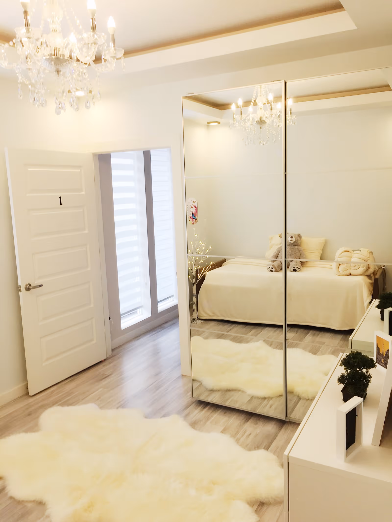A bright bedroom with light wood flooring, a white fluffy rug, a mirrored wardrobe reflecting a bed with cream bedding and a teddy bear, a white door labeled with the number 1, a chandelier hanging from the ceiling, and a white dresser with small plants and framed pictures.