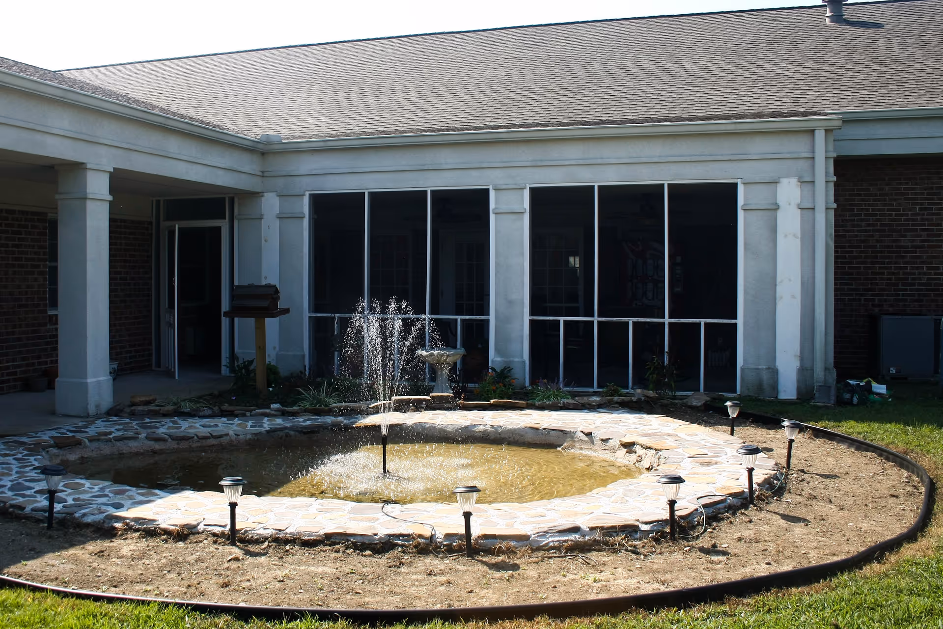 A stone-ringed courtyard fountain with water jets in front of a single-story building with a screened porch and columns.