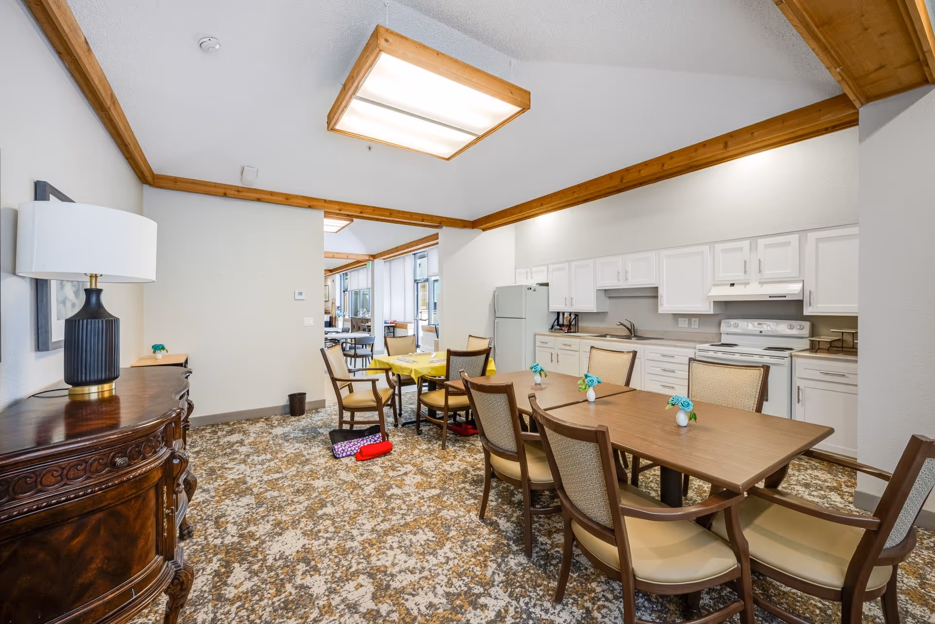 Interior view of a senior living facility dining area with a kitchen in the background. The room features a wooden dining table with six chairs, each with beige cushions. Small vases with blue flowers are placed on the table. The kitchen area has white cabinets, a white refrigerator, stove, and countertop appliances. The room has a patterned carpet, wooden trim along the ceiling, and a decorative wooden sideboard with a lamp on the left side.