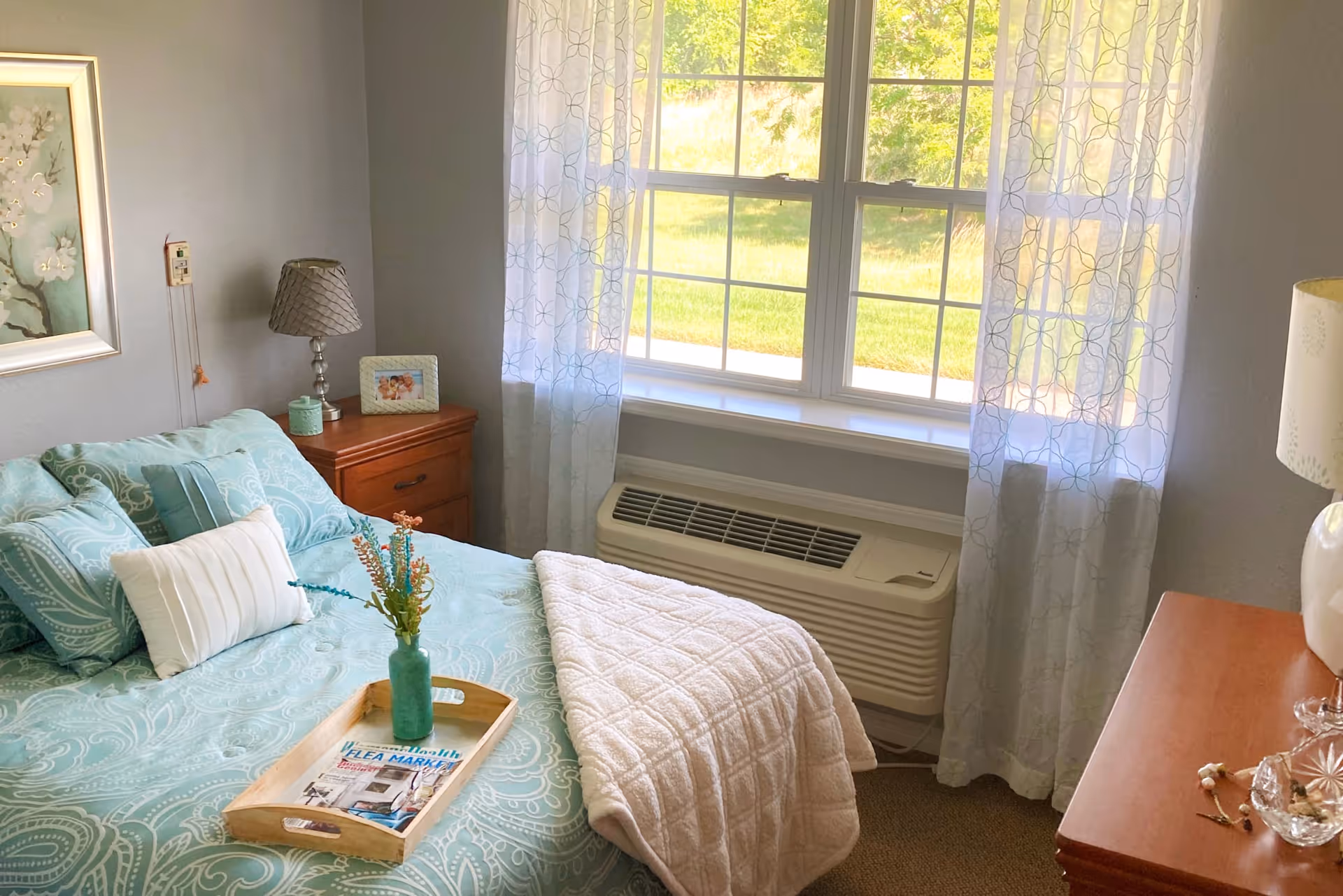 Bright bedroom featuring a teal-patterned bed with decorative pillows, wooden bedside tables, a tray with a vase, and a large window with sheer curtains over a wall air conditioner.