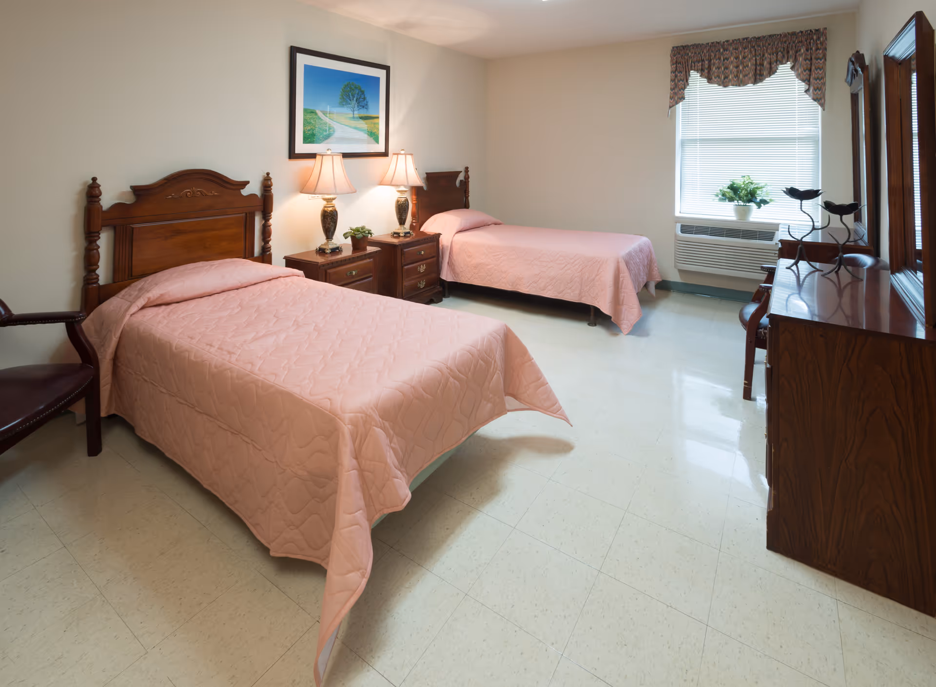 A clean and simple bedroom with two single beds covered in light pink quilts. Between the beds is a wooden nightstand with two table lamps and a small plant. There is a framed picture of a tree on the wall above the nightstand. A window with blinds and a floral valance lets in natural light. On the right side of the room, there is a wooden dresser with decorative items on top and a chair beside it. The floor is tiled and the walls are painted a light beige color.