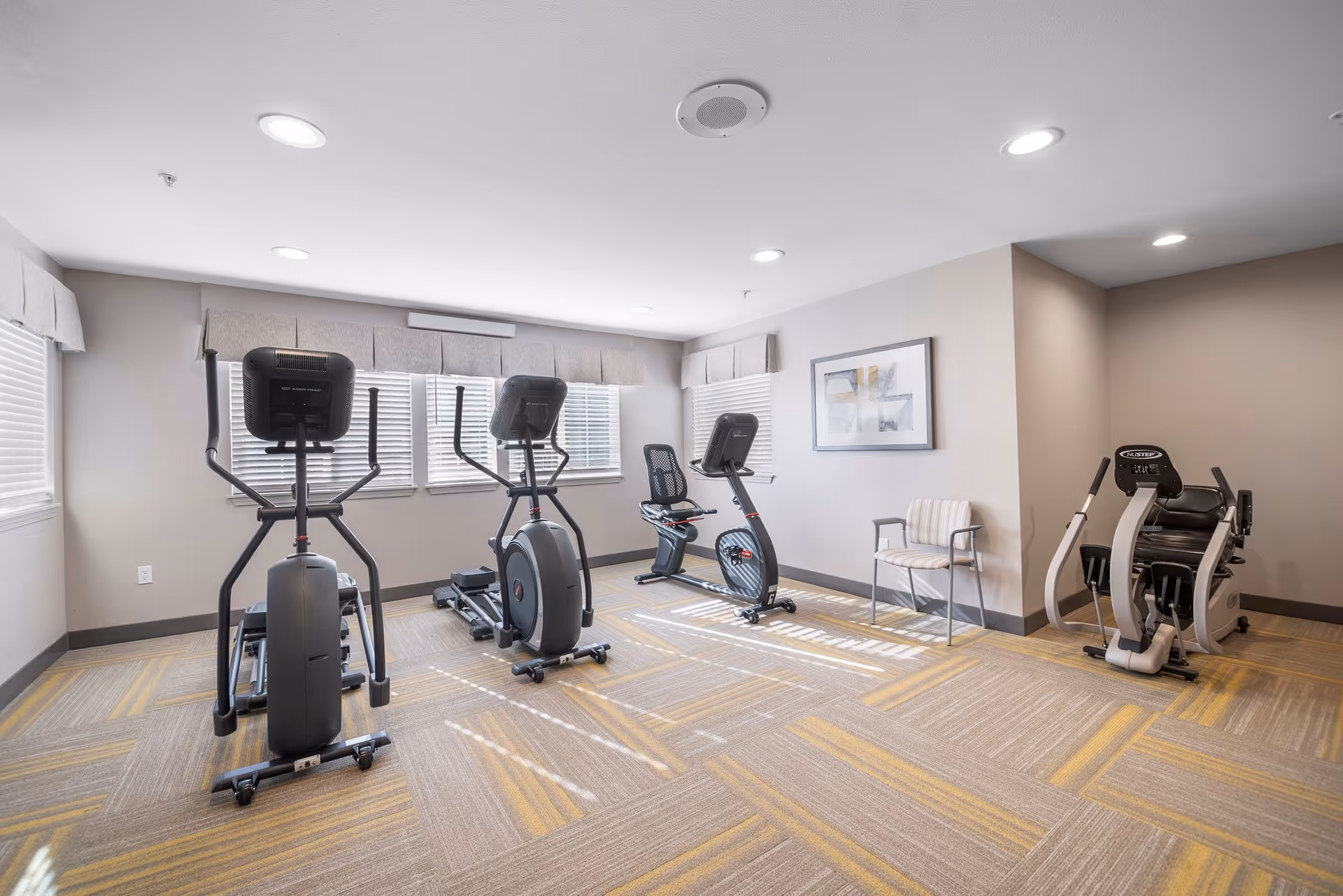 A bright exercise room with three cardio machines including an elliptical, a recumbent bike, and a NuStep machine. The room has beige walls, three windows with blinds and valances, recessed ceiling lights, a framed abstract painting on the wall, and a single striped chair.