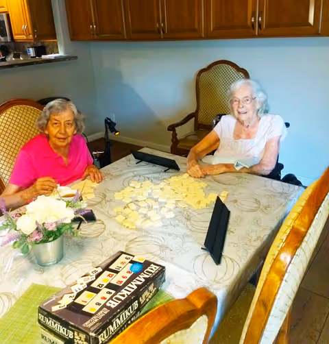 Two elderly women sitting at a dining table playing the game Rummikub. One woman is wearing a pink shirt and the other is in a white top. The table is covered with a patterned tablecloth and has Rummikub tiles spread out, along with game racks and a box. There is a small flower arrangement in a metal pot on the table. The background shows wooden cabinets and chairs.