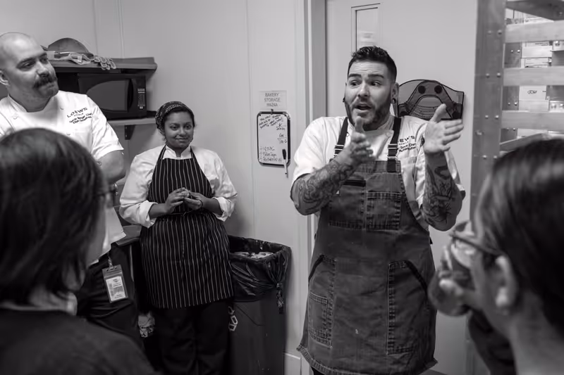 Black and white photo of a group of chefs in a kitchen having a discussion. One chef with tattoos and an apron is speaking and gesturing with his hands, while others listen attentively. The background shows a microwave, a whiteboard with notes, and a trash bin.