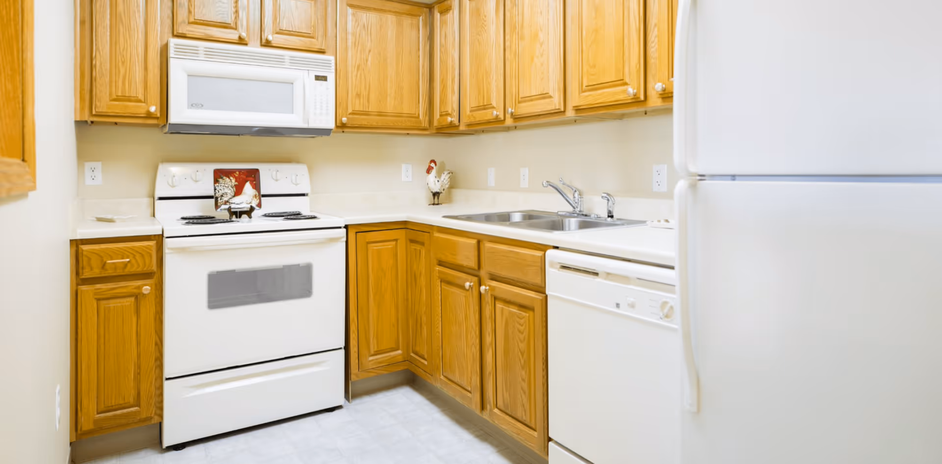 A compact kitchen with wooden cabinets, a white electric stove with a microwave above it, a double sink, a dishwasher, and a refrigerator. The countertops are light-colored, and there is a decorative rooster figurine on the counter.