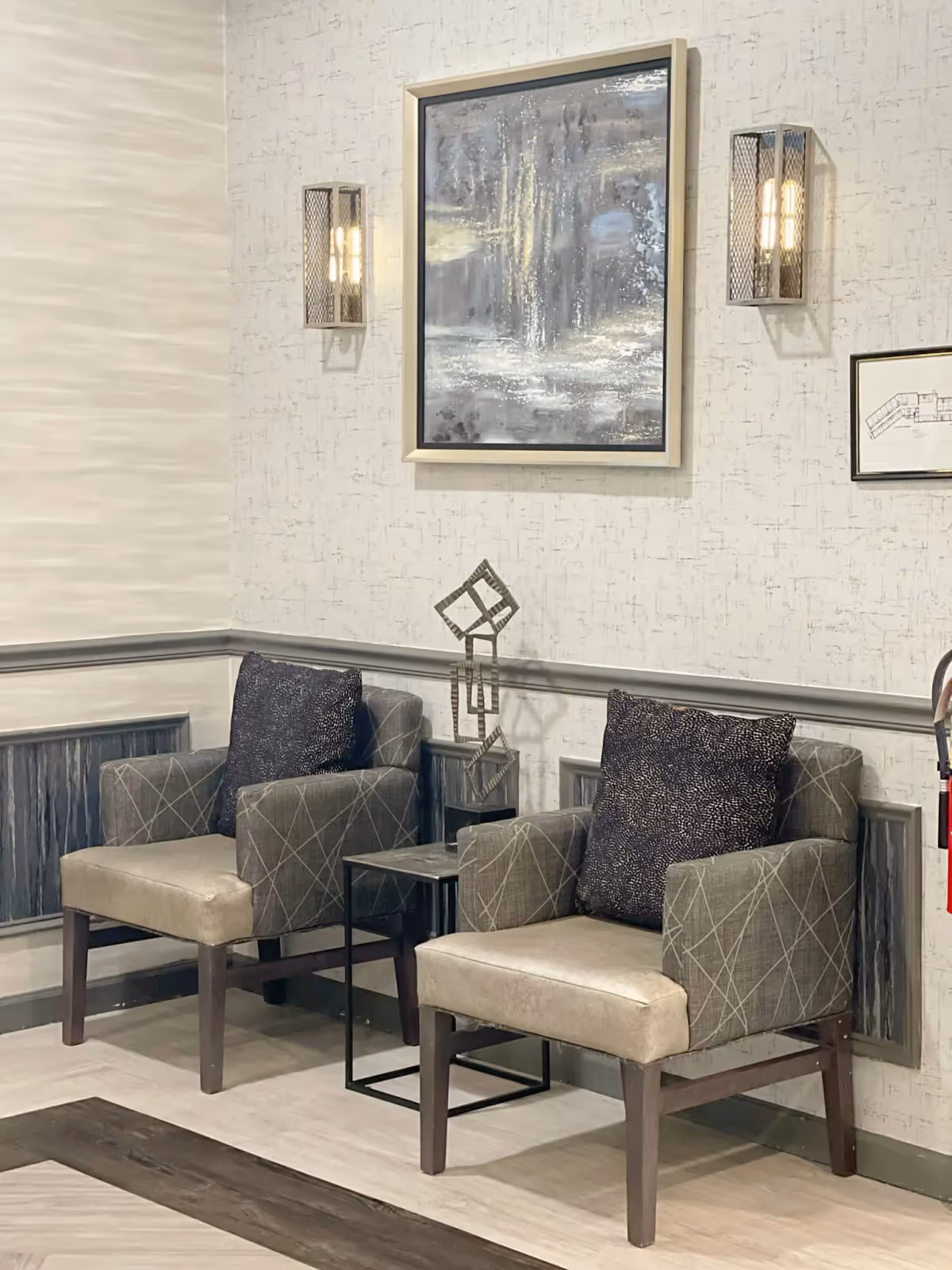 A cozy corner seating area with two cushioned armchairs featuring patterned upholstery and dark throw pillows. Between the chairs is a small black metal side table with a modern geometric sculpture on it. The wall behind has textured wallpaper, two wall sconces with warm lighting, and two framed artworks, one abstract and one architectural drawing.