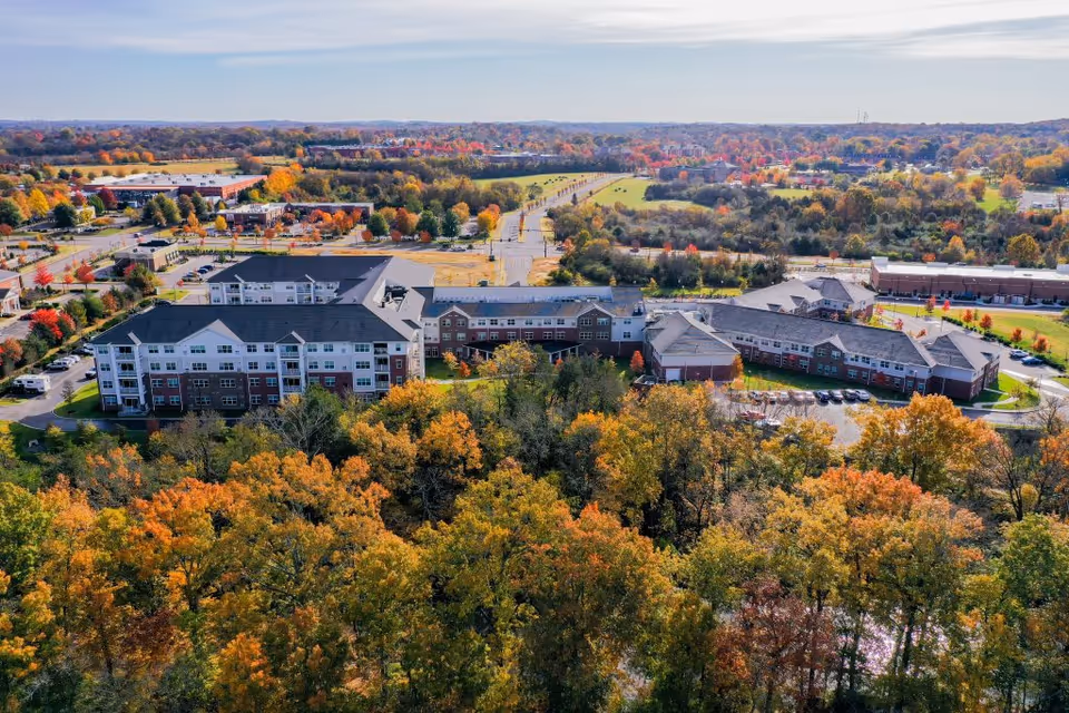 Aerial view of Clarendale at Indian Lake facility surrounded by trees with autumn foliage. The building complex is large with multiple connected sections and parking areas. Roads and other buildings are visible in the background under a clear sky.