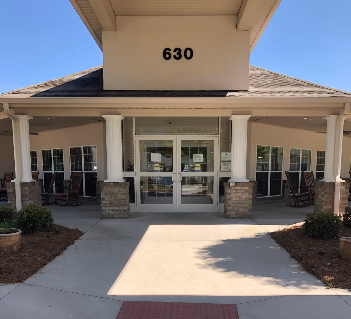 Front entrance of a building with the number 630 above double glass doors and a covered porch with columns and rocking chairs.