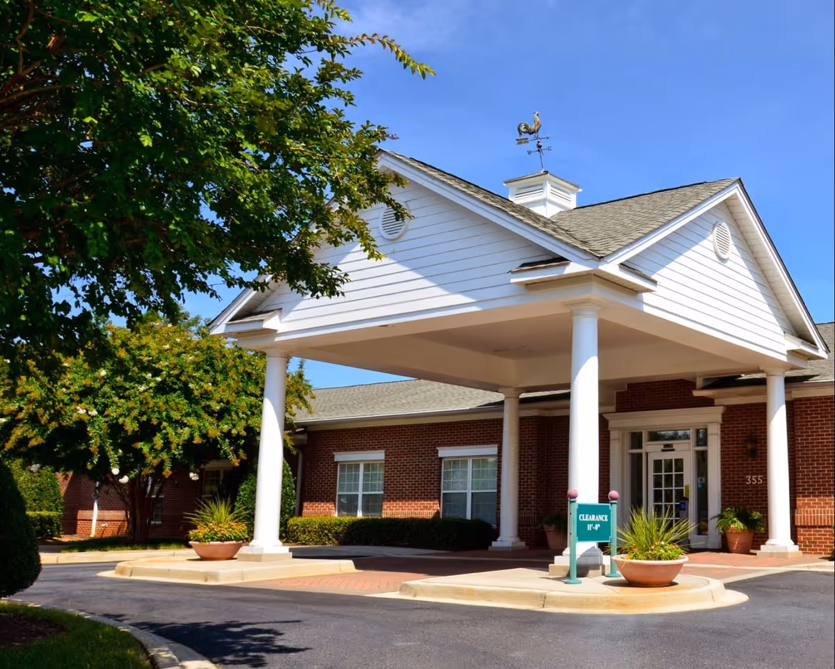 Entrance of a brick building with a white portico supported by four white columns. There is a weather vane on top of the portico roof and a clearance sign indicating 11 feet 4 inches. The building is surrounded by greenery and trees under a clear blue sky.