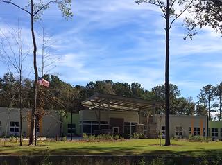 Front exterior of a single-story assisted living building with a covered entrance, flagpole, lawn and trees under a blue sky.