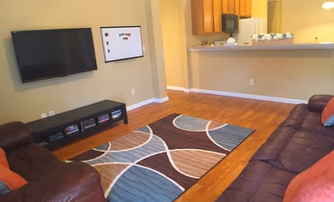 Interior view of a living room with wooden flooring, a patterned area rug, two leather sofas with orange pillows, a wall-mounted flat screen TV, and a whiteboard on the wall. In the background, there is a kitchen counter with wooden cabinets and a microwave.