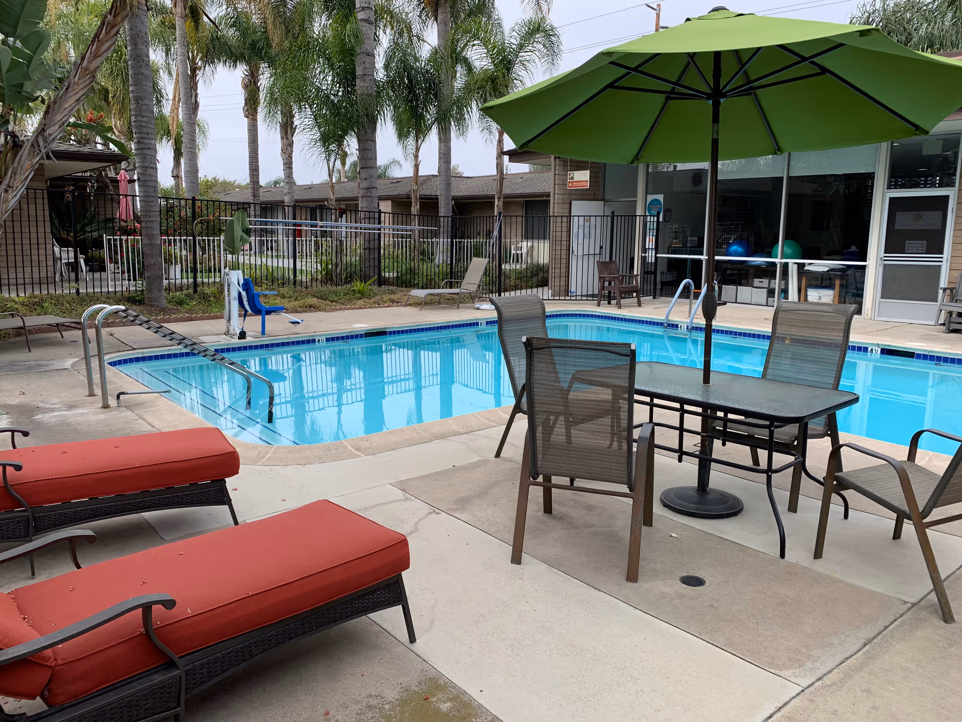 Outdoor swimming pool area with lounge chairs, a table and green umbrella, and palm trees in the background.