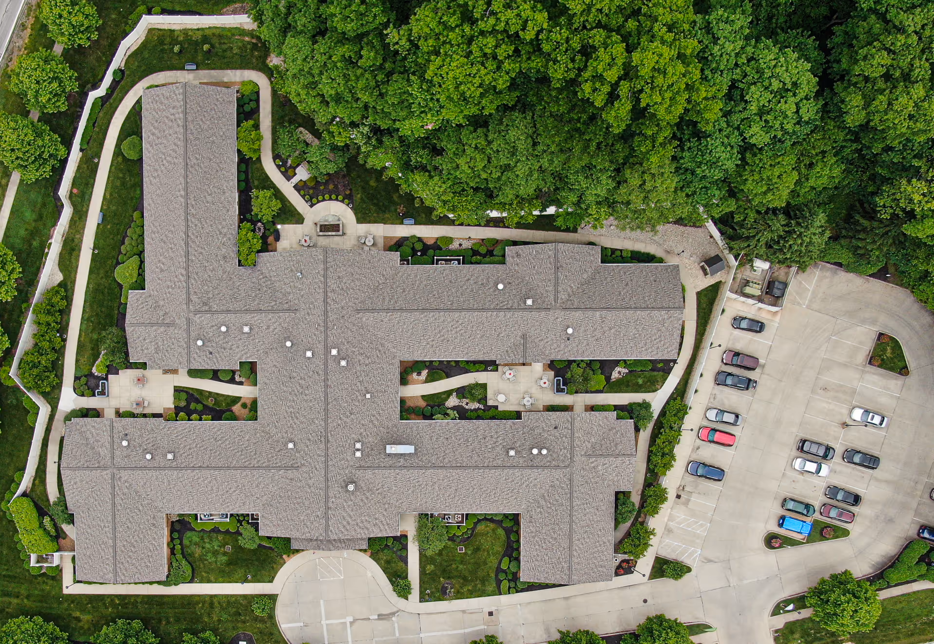 Aerial view of a large H-shaped senior living facility building with surrounding parking, walkways, and dense trees.